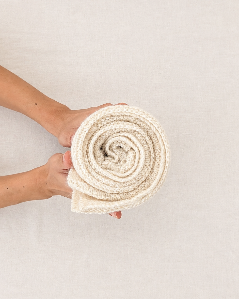 Las manos de una persona sosteniendo una manta de punto enrollada de color crema sobre un fondo neutro de textura beige claro.