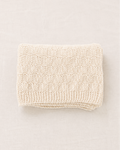 Una manta de punto de color crema, cuidadosamente doblada, con un patrón texturizado de cuadros y un borde de canalé, presentada sobre un fondo neutro de color beige claro.