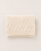 Una manta de punto de color crema, cuidadosamente doblada, con un patrón texturizado de cuadros y un borde de canalé, presentada sobre un fondo neutro de color beige claro.