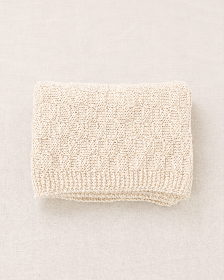 Una manta de punto de color crema, cuidadosamente doblada, con un patrón texturizado de cuadros y un borde de canalé, presentada sobre un fondo neutro de color beige claro.