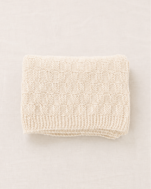 Una manta de punto de color crema, cuidadosamente doblada, con un patrón texturizado de cuadros y un borde de canalé, presentada sobre un fondo neutro de color beige claro.